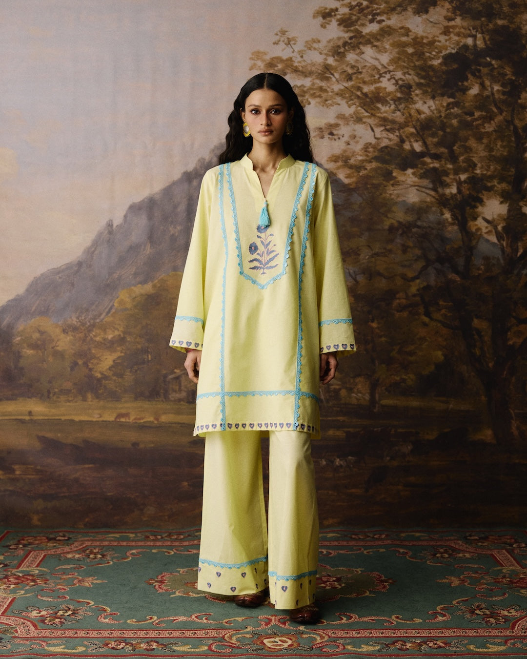 A woman stands in front of a painted landscape backdrop wearing a pale yellow traditional tunic and wide-leg pants set with light blue trim and embroidered detailing. She is also wearing a turquoise pendant necklace.