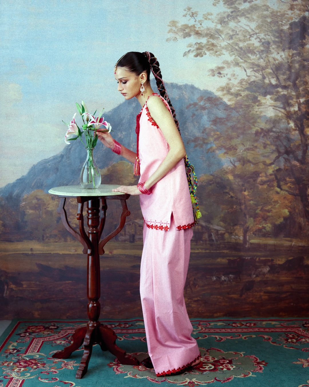 A woman in a pink traditional salwar kameez with red trim stands beside a small round table holding a vase of flowers, set against a painted landscape backdrop.
