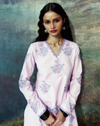 A woman modeling a light pink long-sleeve kurta with purple and green floral embroidery, set against a textured, painterly blue-green background.
