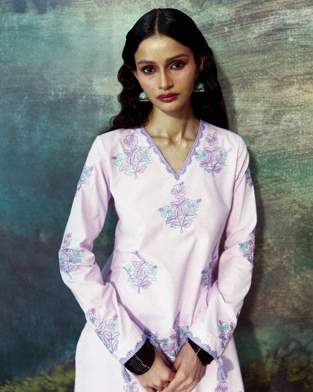 A woman modeling a light pink long-sleeve kurta with purple and green floral embroidery, set against a textured, painterly blue-green background.