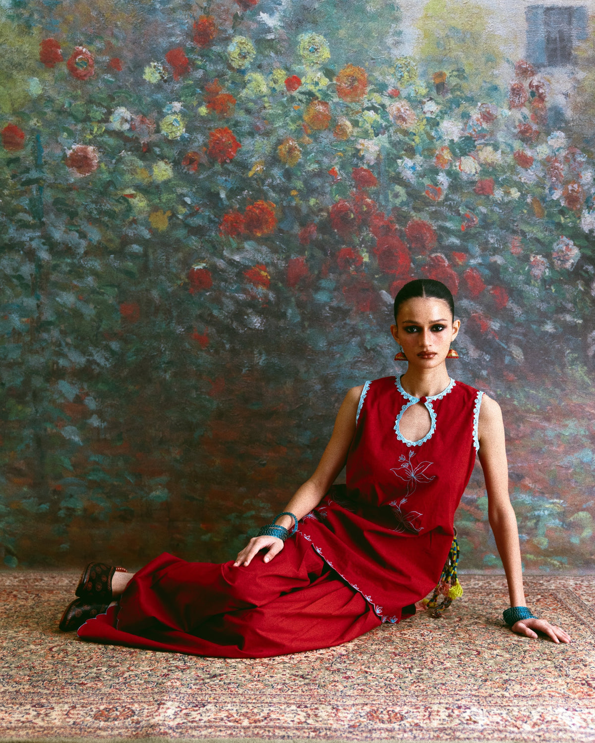 A woman wearing a sleeveless red dress with turquoise trim and jewelry sits on a patterned rug in front of a large floral painting.