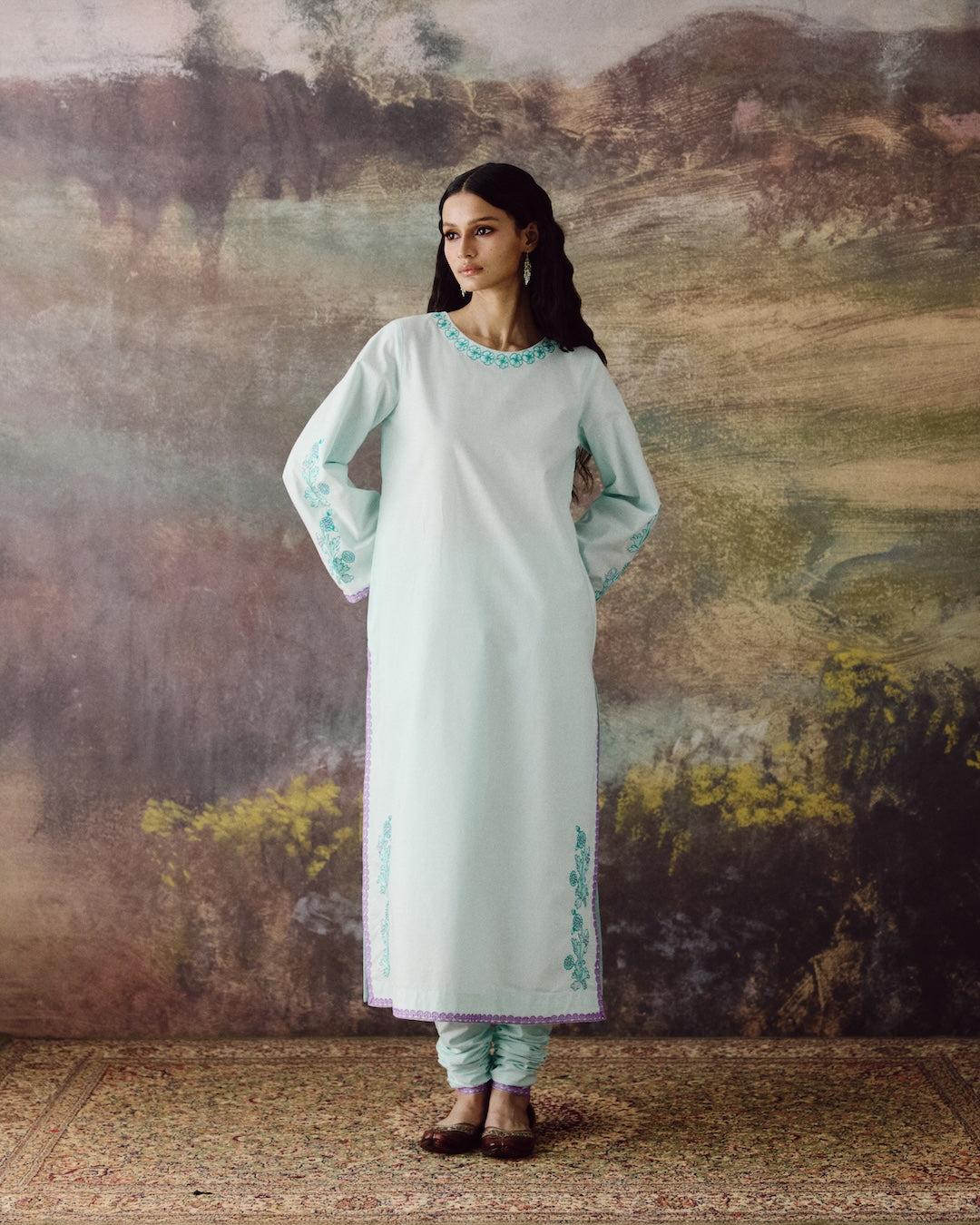 A woman stands in front of a rustic painted backdrop wearing a long mint green kurta with turquoise embroidery, paired with matching pants and brown shoes.
