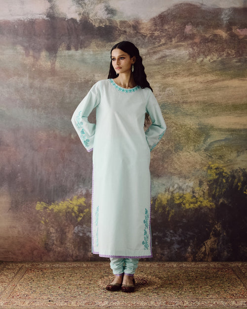 A woman stands in front of a rustic painted backdrop wearing a long mint green kurta with turquoise embroidery, paired with matching pants and brown shoes.