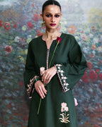 A woman models a dark green long-sleeved tunic dress with pink and white floral embroidery on the cuffs and hem, holding a single red rose against a painterly floral background.