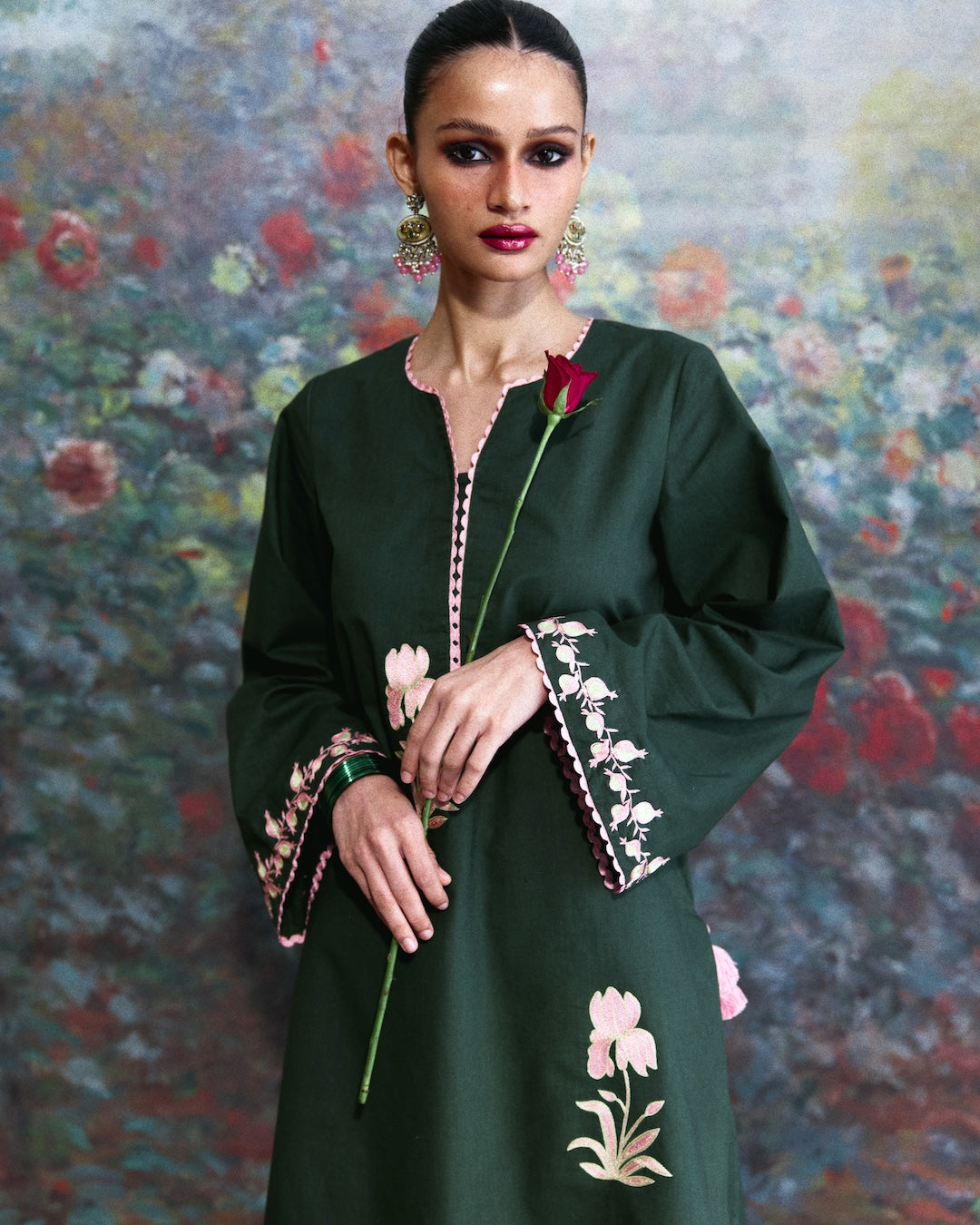 A woman models a dark green long-sleeved tunic dress with pink and white floral embroidery on the cuffs and hem, holding a single red rose against a painterly floral background.