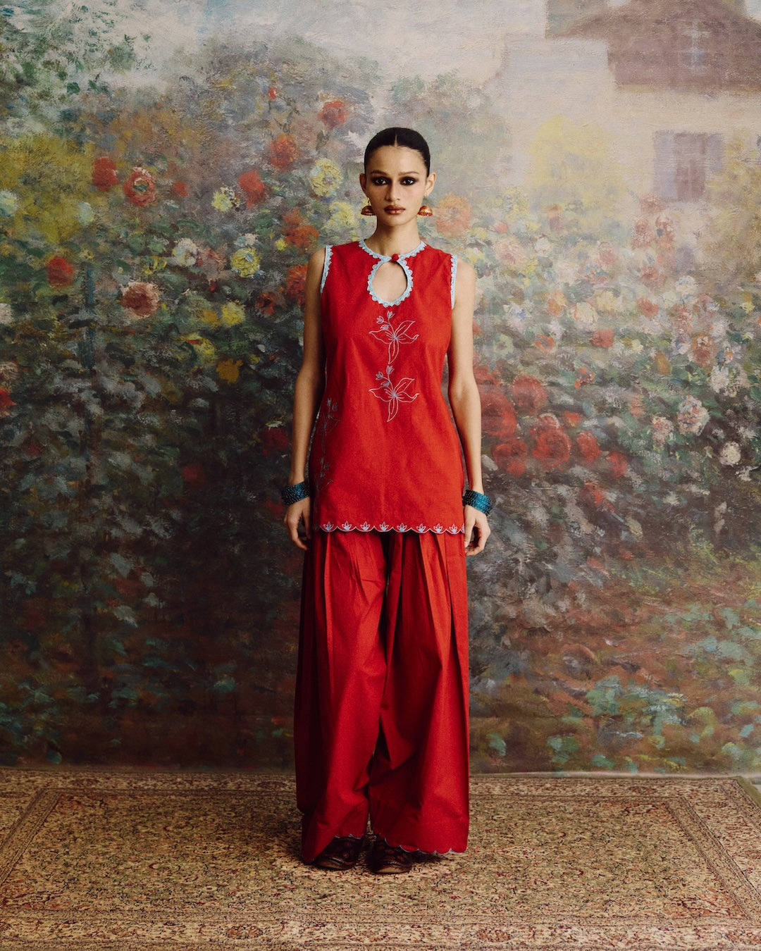A woman stands facing forward in a red sleeveless tunic and matching wide-leg pants set, with small white embroidered floral details and a silver-toned keyhole neckline. She is posed in front of a large painted floral wall and a patterned rug.