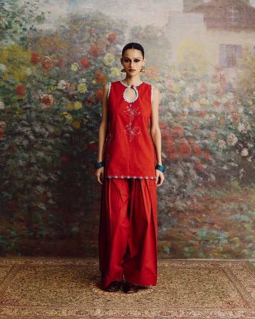 A woman stands facing forward in a red sleeveless tunic and matching wide-leg pants set, with small white embroidered floral details and a silver-toned keyhole neckline. She is posed in front of a large painted floral wall and a patterned rug.