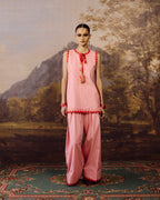 A model stands in front of a painted landscape backdrop wearing a light pink traditional outfit with red embroidered trim and a matching pink dupatta. The setting includes a patterned rug and a scenic painting in the background.