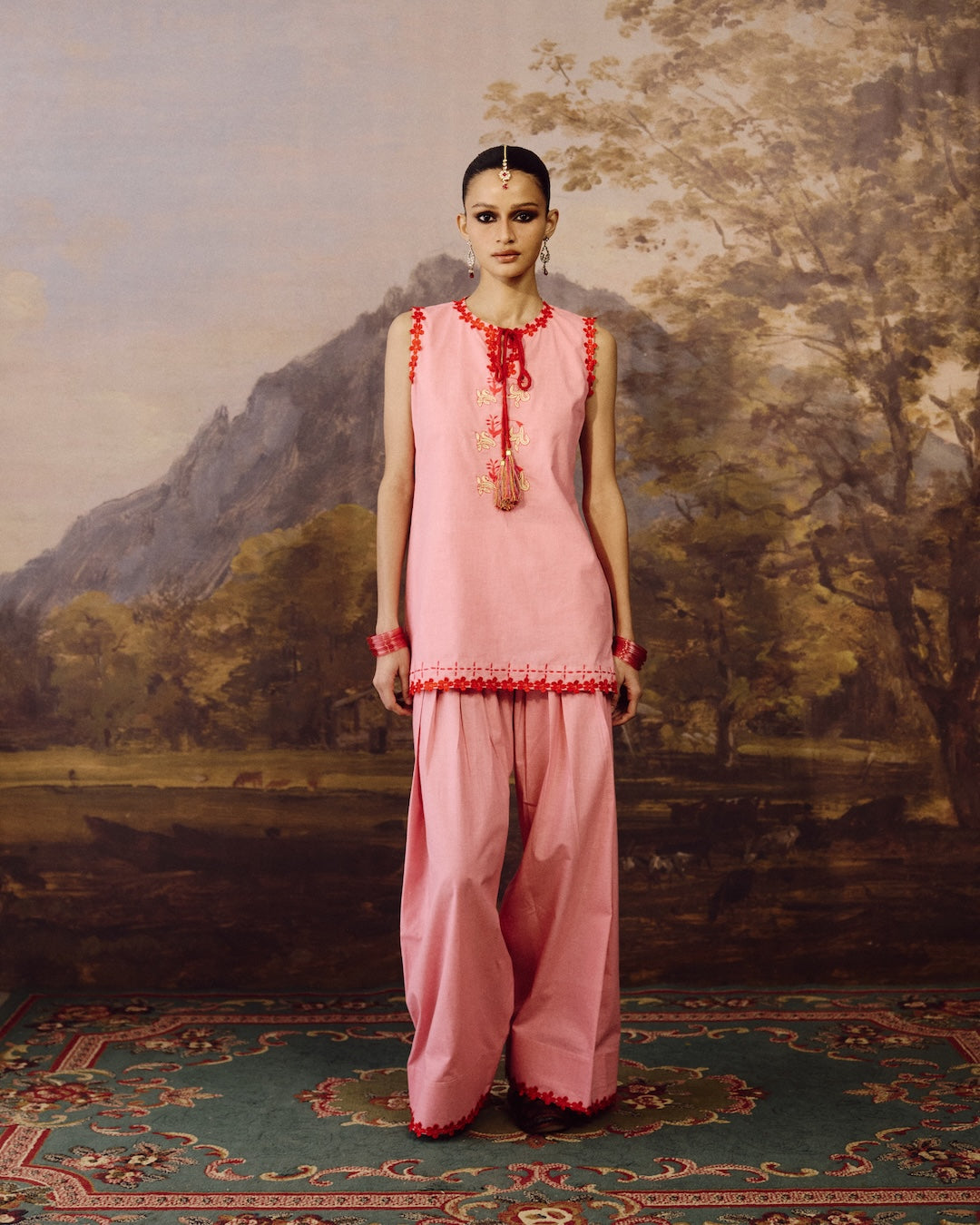 A model stands in front of a painted landscape backdrop wearing a light pink traditional outfit with red embroidered trim and a matching pink dupatta. The setting includes a patterned rug and a scenic painting in the background.