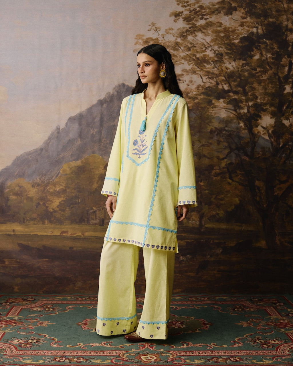A woman modeling a light yellow traditional outfit with light blue embroidery and geometric borders, standing on a patterned rug in front of a painted landscape backdrop.