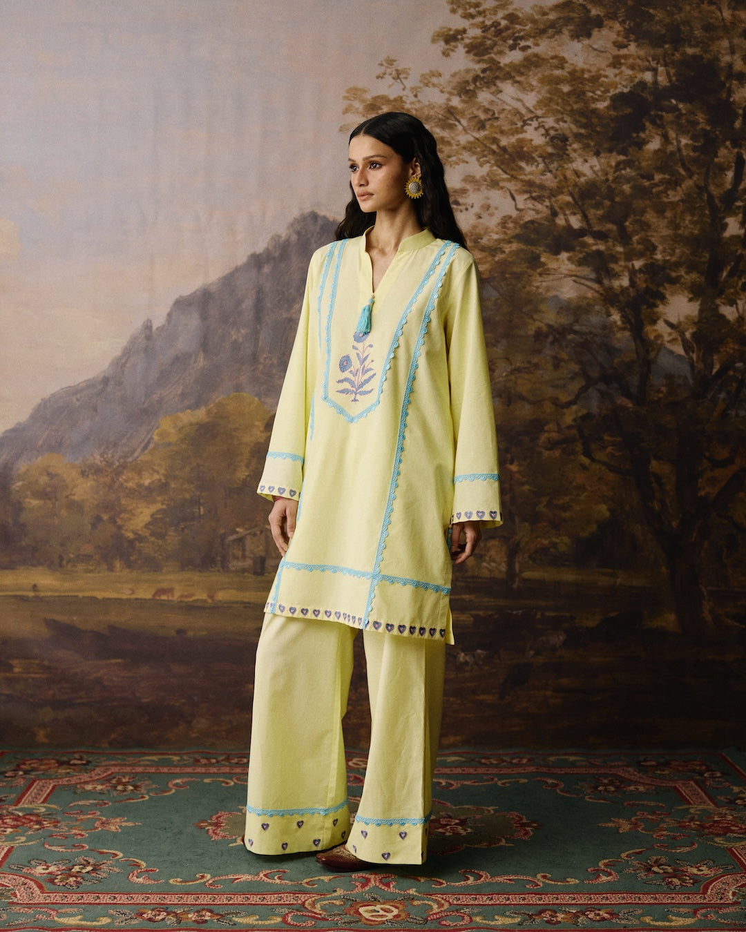 A woman modeling a light yellow traditional outfit with light blue embroidery and geometric borders, standing on a patterned rug in front of a painted landscape backdrop.