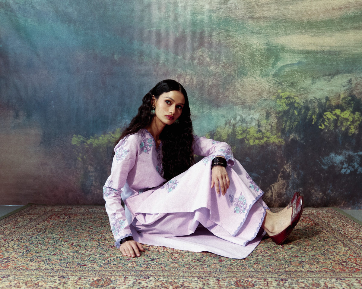 A woman sits cross-legged on a patterned rug in front of a textured, painterly backdrop, wearing a lavender traditional outfit with floral embroidery and dark red flat shoes.