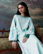 A woman seated on a patterned upholstered bench wearing a long, flowing mint green tunic with turquoise floral embroidery around the neckline and on the sleeves.