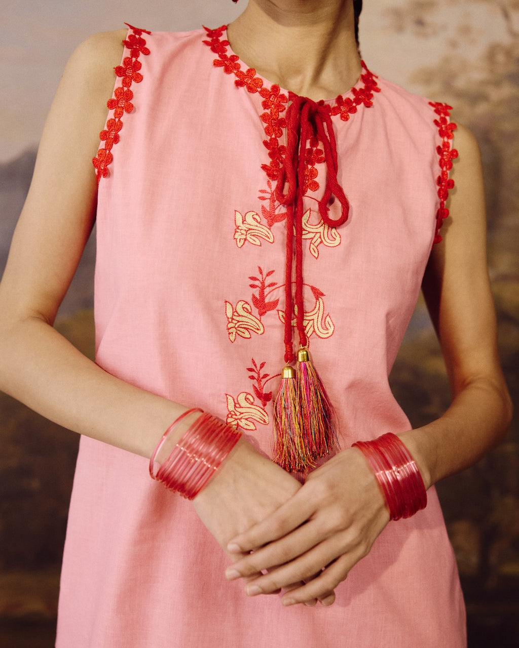A close-up of a model wearing a sleeveless pink dress with red floral embroidery along the neckline and armholes, red tassel tie details, and gold floral embroidery on the front. The model is also wearing red bangles.