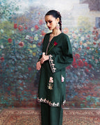 A woman models a dark green embroidered kurta set with matching trousers, standing in front of a painted floral background. The kurta features pink and white floral embroidery on the front, sleeve cuffs, and hem.