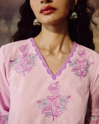 A close-up of a woman wearing a light pink embroidered top with purple and light blue floral embroidery and a purple lace-trimmed V-neckline. She is wearing ornate gold-toned earrings, and her dark hair is visible against a softly blurred warm-toned background.