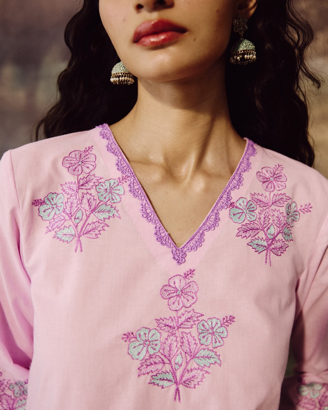 A close-up of a woman wearing a light pink embroidered top with purple and light blue floral embroidery and a purple lace-trimmed V-neckline. She is wearing ornate gold-toned earrings, and her dark hair is visible against a softly blurred warm-toned background.