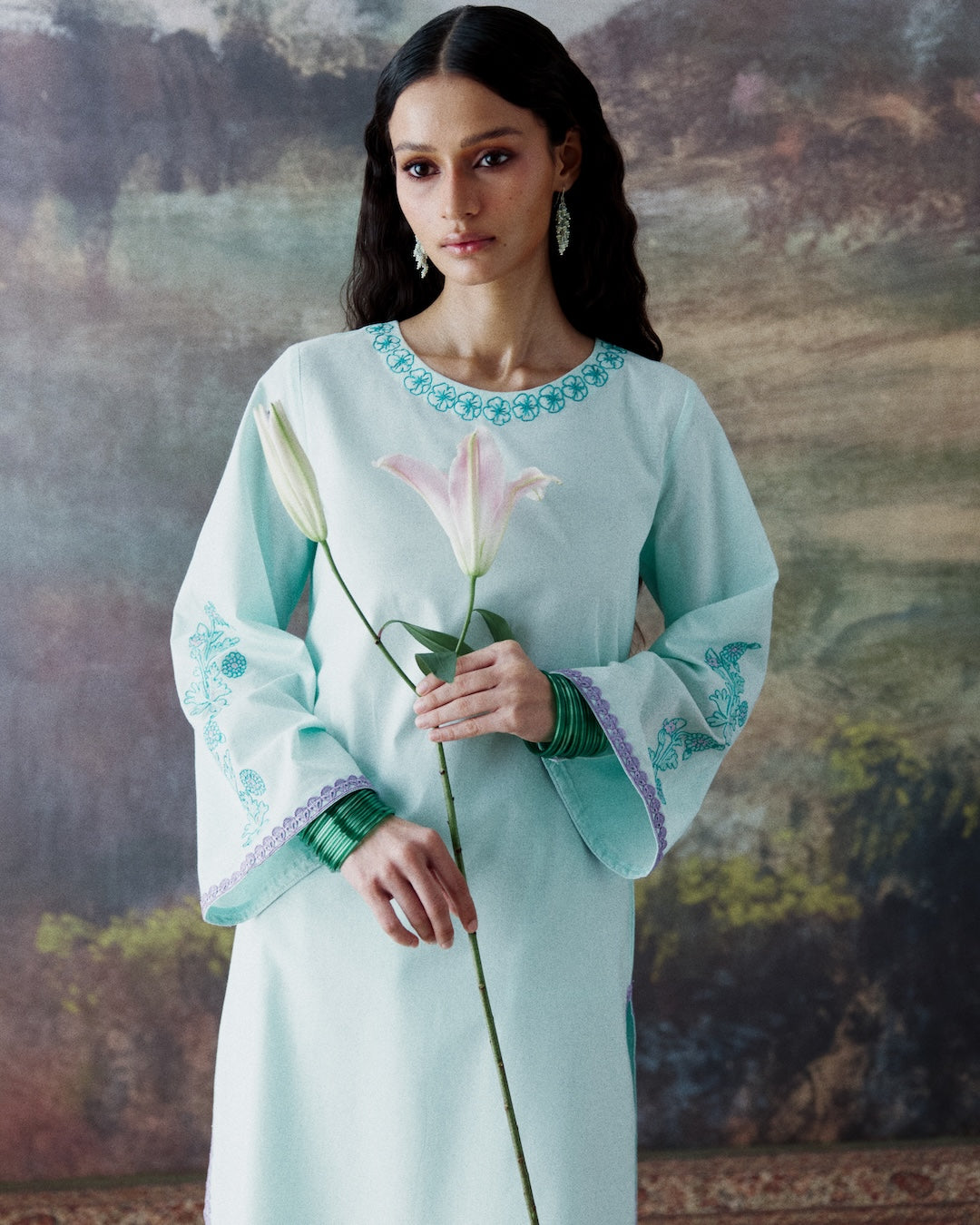A woman modeling a light mint green traditional long-sleeve dress with blue and purple embroidery, holding a pink lily flower against a scenic painted backdrop.