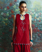 A woman modeling a sleeveless red kurta with light blue and white embroidery, paired with matching red flared pants, set against a painterly floral background.