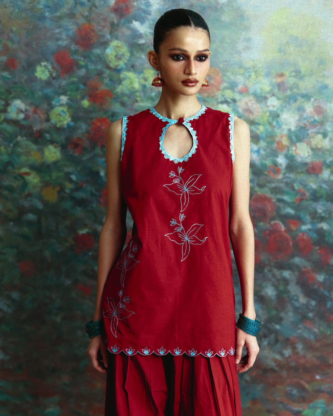 A woman modeling a sleeveless red kurta with light blue and white embroidery, paired with matching red flared pants, set against a painterly floral background.
