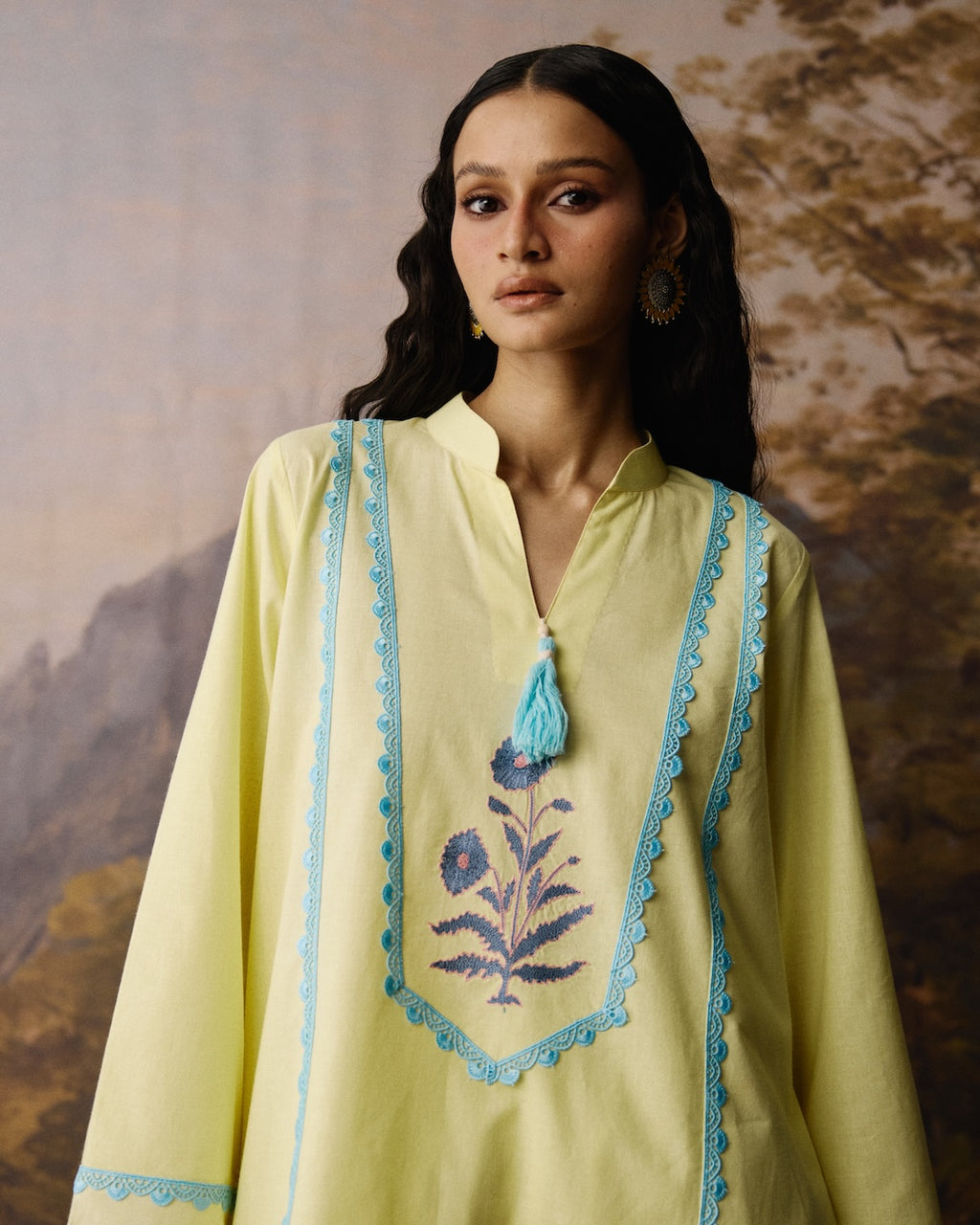 A woman wearing a pale yellow tunic with light blue embroidered borders, a blue tassel, and a blue floral motif on the chest, posed against a textured, warm-toned painted backdrop.