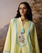 A woman wearing a pale yellow tunic with light blue embroidered borders, a blue tassel, and a blue floral motif on the chest, posed against a textured, warm-toned painted backdrop.