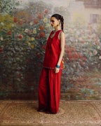 A woman stands in profile wearing a red sleeveless tunic and matching wide-leg pants, posed in front of a large painted floral backdrop with a patterned rug beneath her.