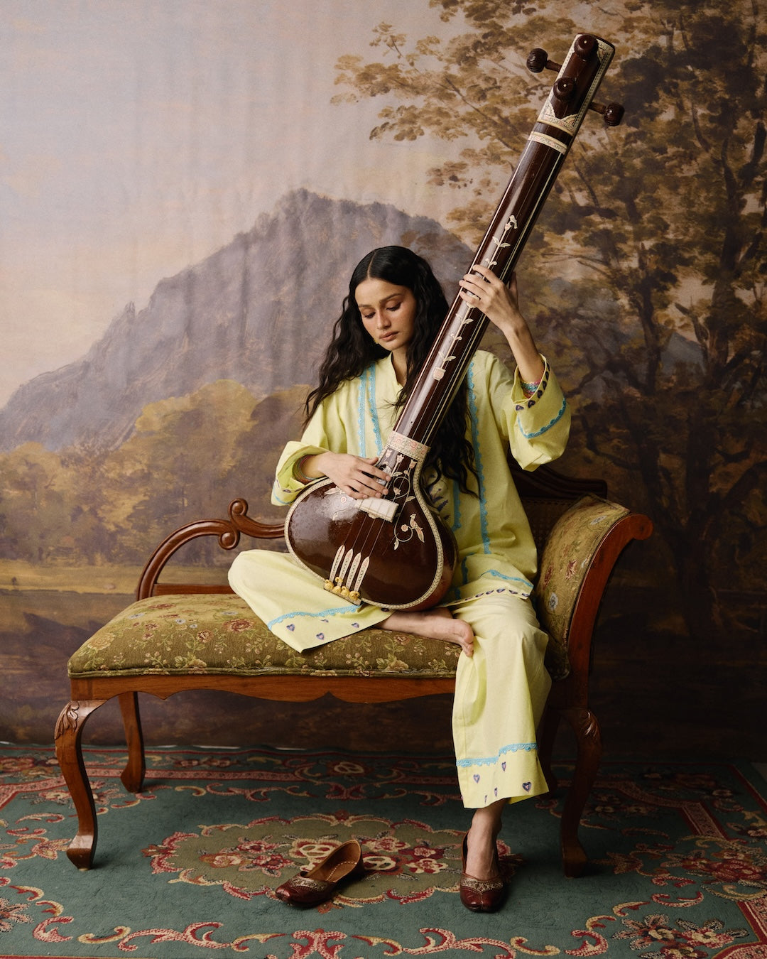 A woman sits barefoot on an ornate vintage chaise lounge, holding and playing a traditional Indian string instrument called a veena. She is wearing a light yellow-green kurta with blue embroidery.