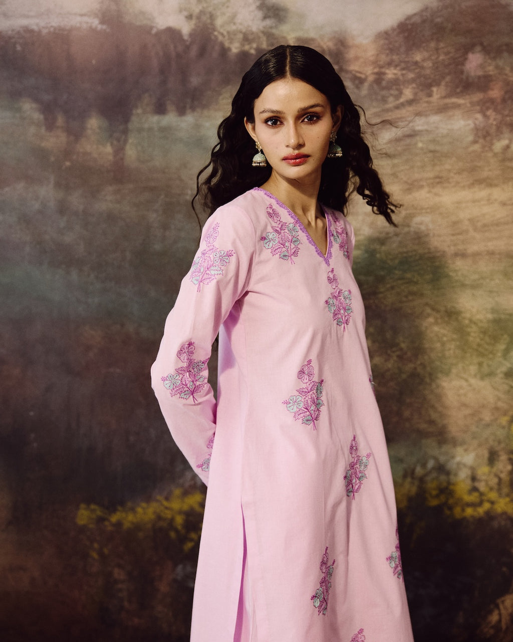 A woman wearing a light pink long kurta with pink and purple floral embroidery, posed against a painterly, scenic background.