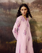 A woman wearing a light pink long kurta with pink and purple floral embroidery, posed against a painterly, scenic background.