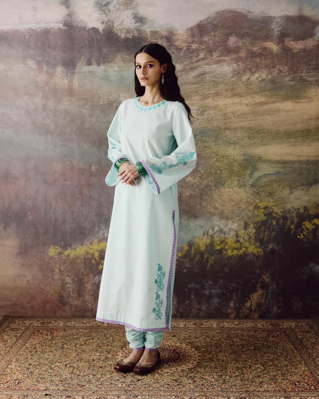 A woman modeling a light mint green kurta with teal embroidery and purple border details, paired with matching pants, standing in front of a rustic, textured landscape backdrop.