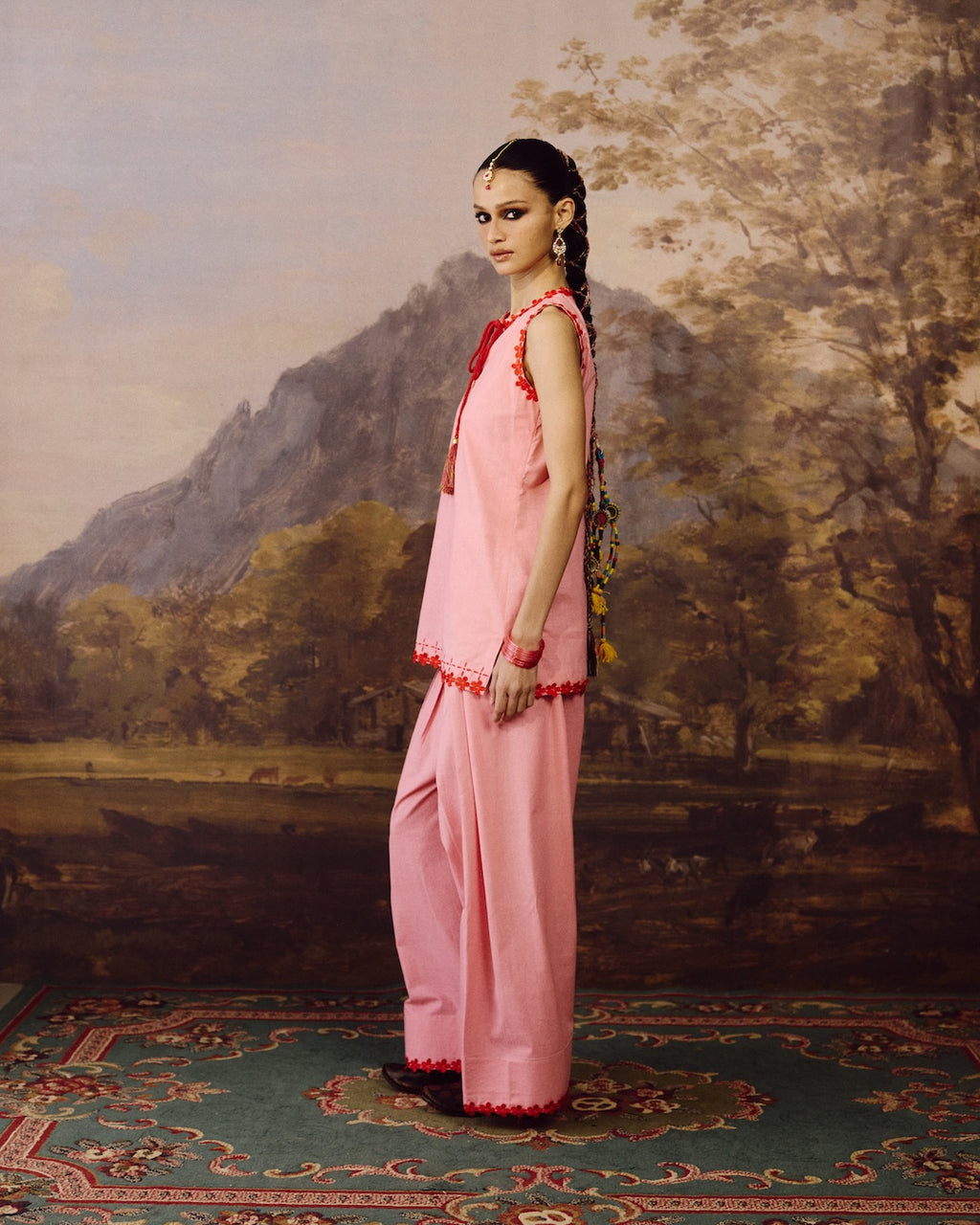 A model stands in profile wearing a light pink traditional outfit with red embroidered trim, posed against a classical landscape painting backdrop of mountains and trees.
