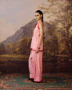 A model stands in profile wearing a light pink traditional outfit with red embroidered trim, posed against a classical landscape painting backdrop of mountains and trees.
