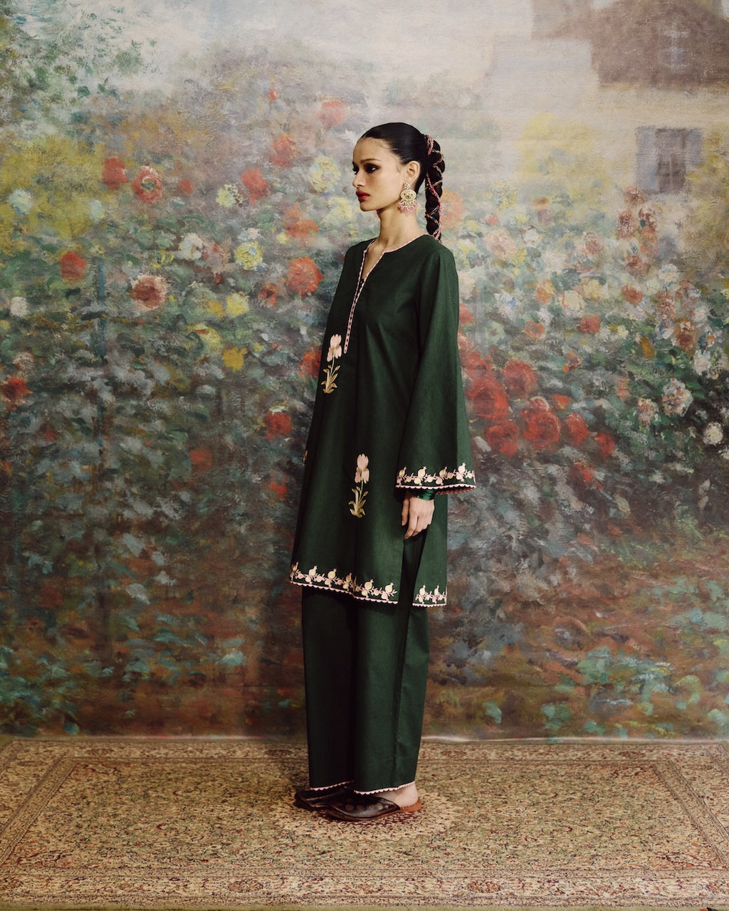 A woman stands in profile wearing a dark green traditional outfit with embroidered borders, posed against a large floral wall mural and a patterned rug.
