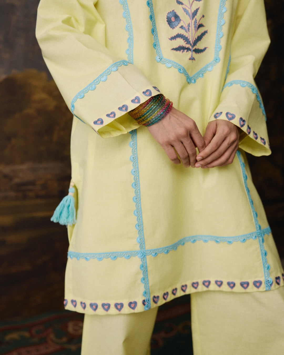 A close-up of a person wearing a pale yellow embroidered kurta with light blue border trim, purple heart motifs, and a matching light yellow trouser. The kurta features floral embroidery on the chest and a tassel detail on the sleeve.