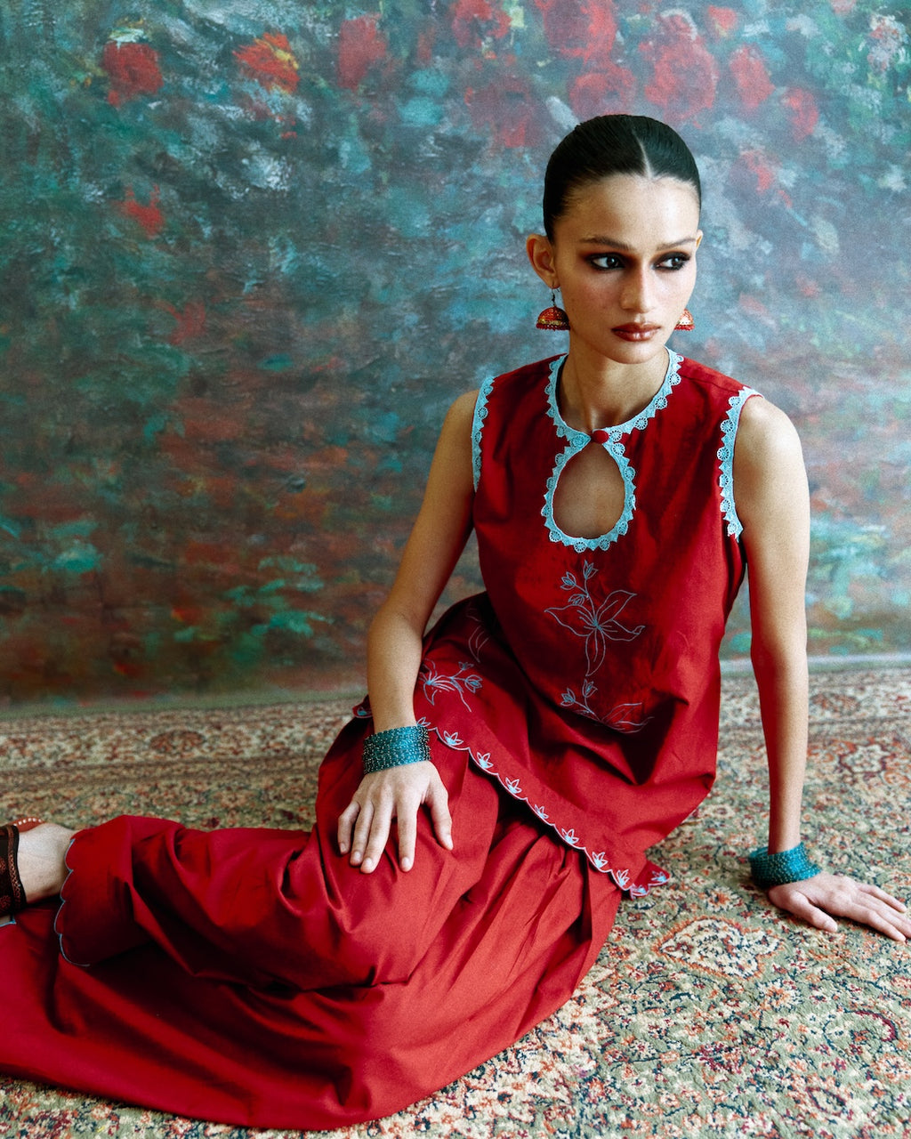 A woman wearing a red sleeveless kurta with light blue lace trim and matching red pants, sitting on a patterned rug against a textured painted backdrop.