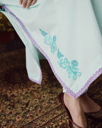 A close-up of a light mint green or pale green fabric garment with turquoise floral embroidery and a lavender scalloped border, worn by a person seated on a patterned rug.