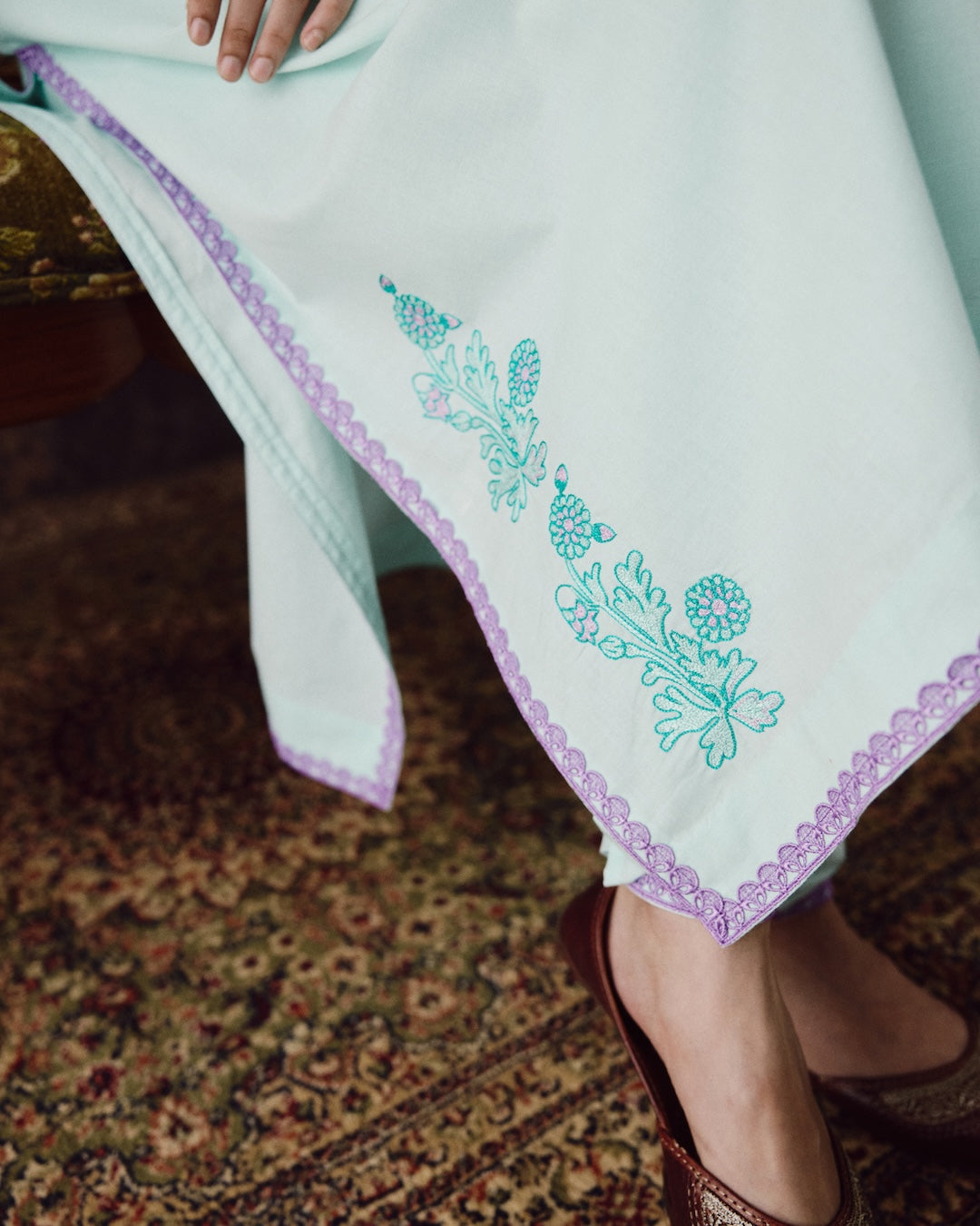 A close-up of a light mint green or pale green fabric garment with turquoise floral embroidery and a lavender scalloped border, worn by a person seated on a patterned rug.