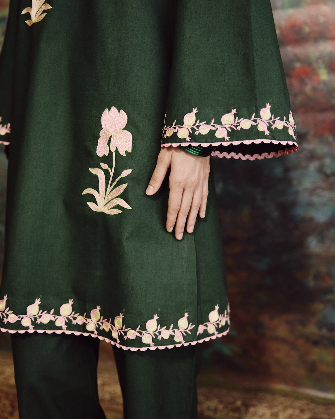 A close-up of a dark green traditional outfit with pink and beige floral embroidery and scalloped borders, worn by a model whose hand is visible.