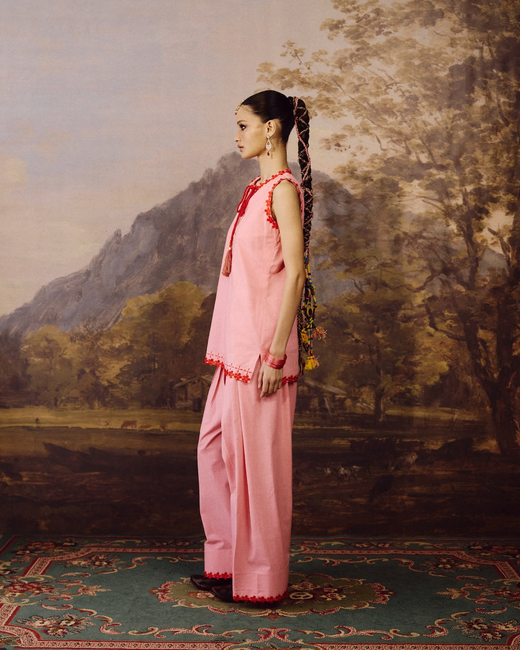A woman stands in profile wearing a light pink traditional outfit with red embroidered trim, posed in front of a painted landscape backdrop featuring mountains, trees, and a patterned rug.