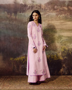 A woman stands in front of a painted landscape backdrop wearing a long light pink traditional outfit with floral embroidery in pink and purple tones. The outfit includes a long kurta-style top and matching wide-leg pants.