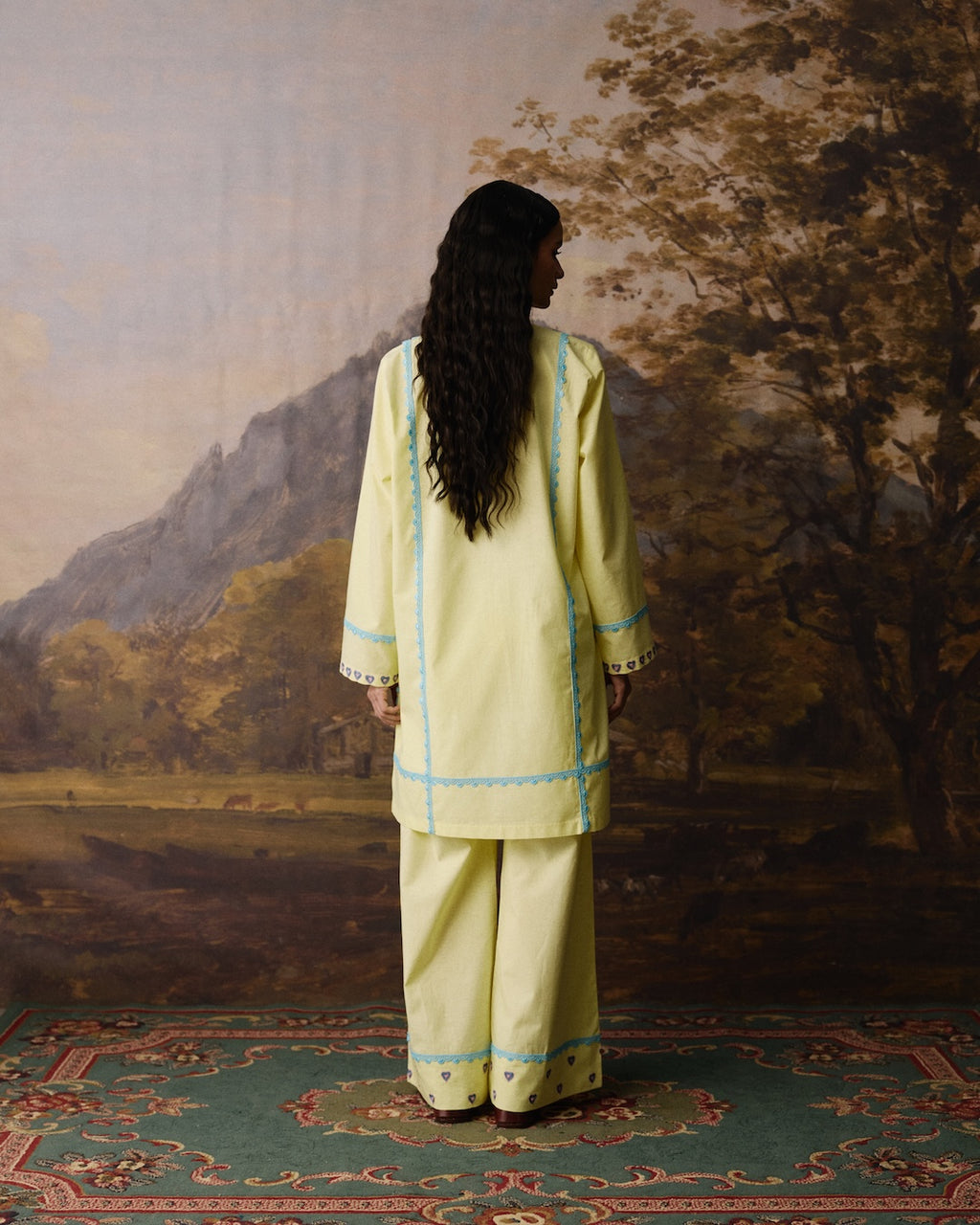 A woman stands with her back to the camera wearing a light yellow-green traditional kurta and trouser set with light blue embroidered borders, posed in front of a scenic landscape painting.
