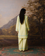 A woman stands with her back to the camera wearing a light yellow-green traditional kurta and trouser set with light blue embroidered borders, posed in front of a scenic landscape painting.