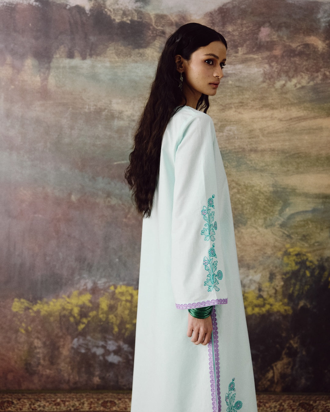 A woman wearing a long white kaftan dress with teal floral embroidery on the sleeve and a purple lace trim, standing in front of a textured, painterly background.