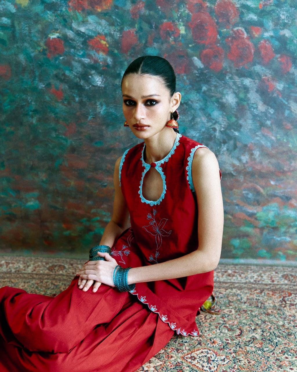 A woman seated on a patterned rug in front of a textured, painterly backdrop. She is wearing a sleeveless red dress with light blue embroidered trim around the neckline and hem, along with blue bracelets.