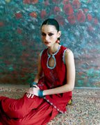 A woman seated on a patterned rug in front of a textured, painterly backdrop. She is wearing a sleeveless red dress with light blue embroidered trim around the neckline and hem, along with blue bracelets.