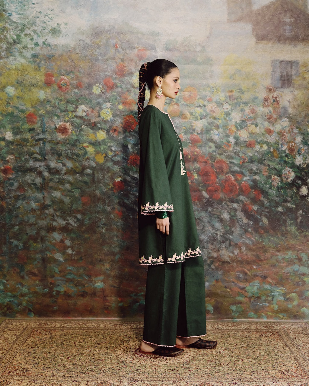 A woman stands in profile wearing a dark green traditional outfit with embroidered details near the cuffs and hem, posed against a large floral painting on a wall.