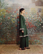 A woman stands in profile wearing a dark green traditional outfit with embroidered details near the cuffs and hem, posed against a large floral painting on a wall.