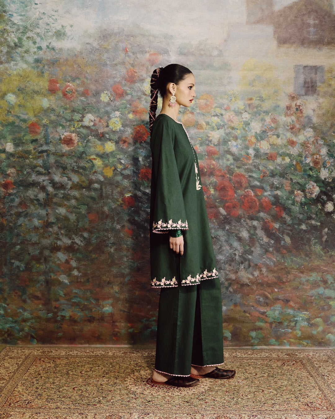 A woman stands in profile wearing a dark green traditional outfit with embroidered details near the cuffs and hem, posed against a large floral painting on a wall.