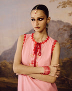A woman modeling a pink sleeveless traditional-style top with red embroidered trim and a red tie neckline, posed against a soft painted landscape backdrop.